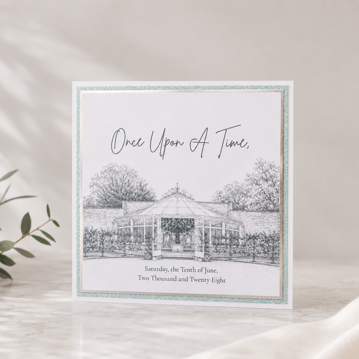 A square wedding invitation featuring a black-and-white sketch of the glasshouse at Combermere Abbey with decorative script text reading 'Once Upon A Time,' displayed upright on a light marble surface with soft shadows and a blurred green branch at the side.