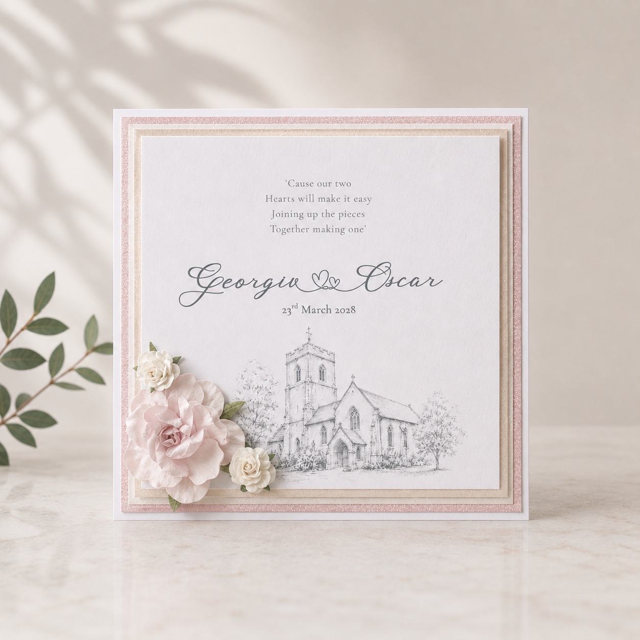 A square wedding invitation card with a church sketch and the names Georgina & Oscar, styled with pale pink and white flowers on a light marble surface with soft leaf shadows.