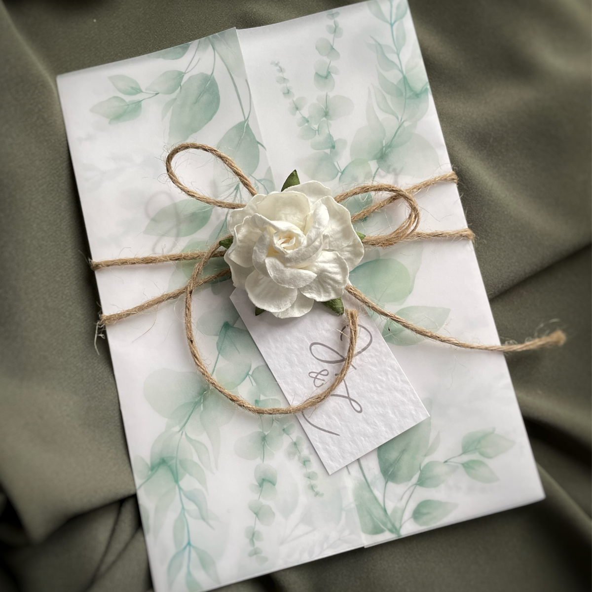 Luxury sage green eucalyptus wedding invitation wrapped in vellum and finished with rustic twine, a handmade white flower and personalised tag.