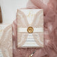 Intricate lace design wedding invite in blush pink shimmer card with gold wax seal and ribbon belly band